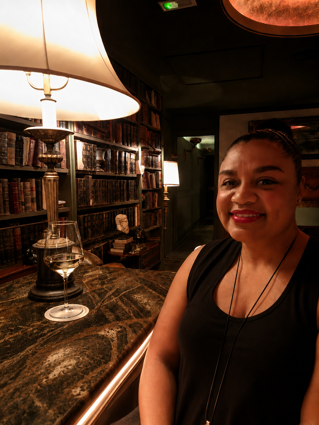 A woman smiling in a warmly lit library lined with old books, beside a glass of white wine