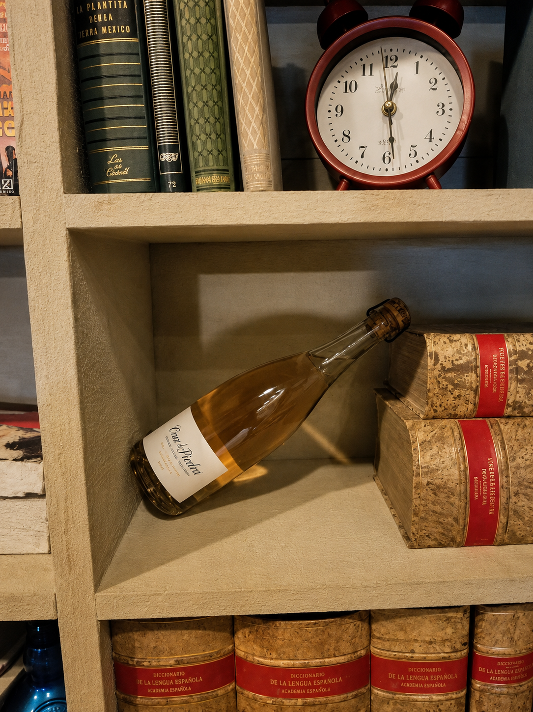 A bottle of white wine lying on a shelf beside books and a red alarm clock